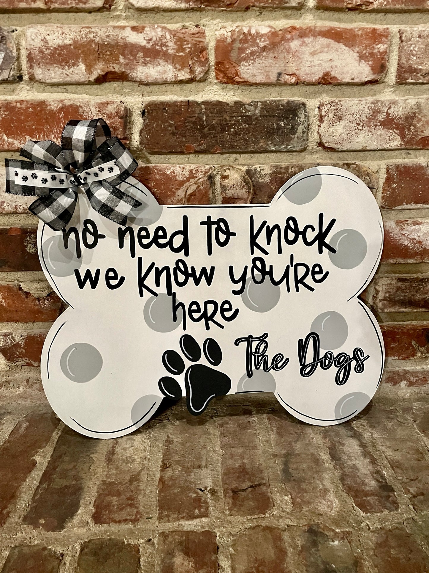 a bone shaped door hanger that reads "no need to knock we know you're here - The Dogs" that's leaning on a brick wall