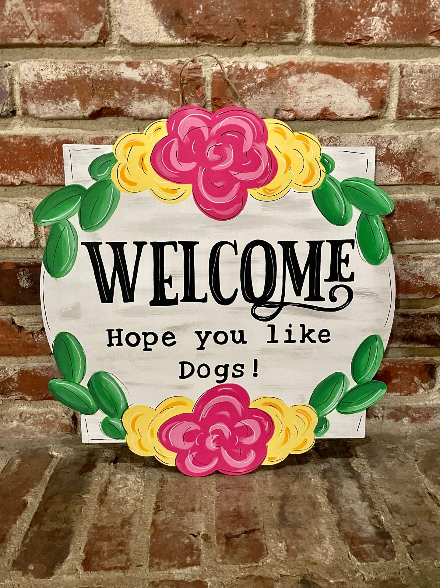 A door hanger that says "Welcome Hope you like dogs" with a floral border leaning against a brick wall