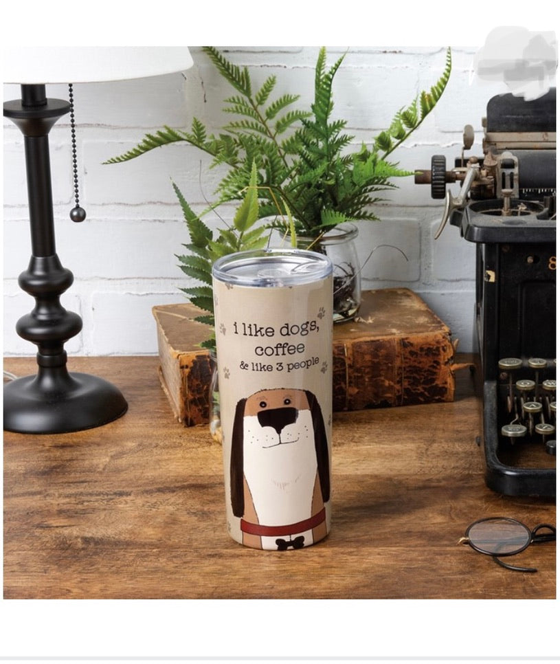 Staged picture of a tan tumbler with paw prints and a dog with the text "I like dogs, coffee, and like 3 people" on a wooden desk with a lamp, book, and plants